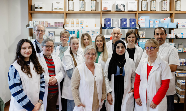 Fröhliche Mitarbeiterin der Gambrinus Apotheke vor Regalen mit Medizinprodukten, repräsentiert Teamgeist und Kundenservice, veranschaulicht die positive Arbeitsatmosphäre und Mitarbeiterzufriedenheit.